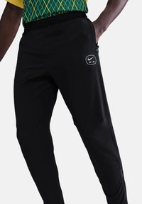 Pantalons de sport noirs en tissu élastique, dotés d'une poche zippée et d'un logo circulaire avec "AIR". Coupe ajustée et texture lisse.