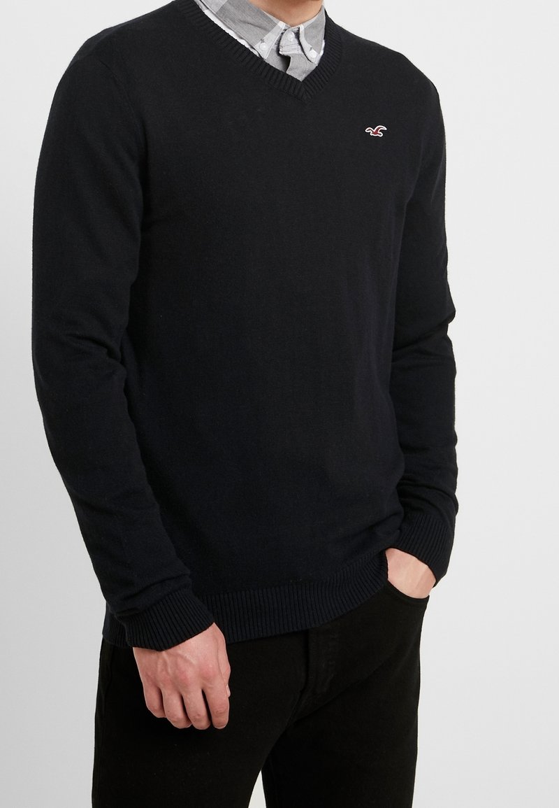 Pull en coton bleu marine avec un col en V et des poignets et ourlet côtelés, arborant un petit logo brodé sur la poitrine. Associé à une chemise à col.