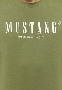 Olivgrön t-shirt i mjuk bomull, med stor vit text: "MUSTANG JEANS COMPANY SINCE 1932" i fet stil.