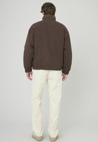 Veste matelassée marron à motif à carreaux, col montant et poignets élastiques, associée à un pantalon ivoire et des chaussures beiges.