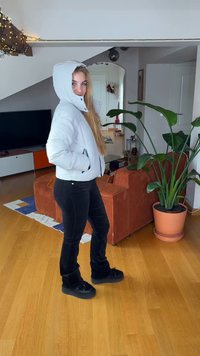 Doudoune à capuche gris clair avec poches latérales, fermetures éclair noires, associée à un pantalon noir ajusté et des chaussures noires épaisses, dans un cadre domestique.