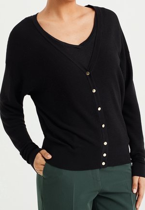 Femme portant un cardigan noir boutonné sur un haut noir avec un pantalon vert, main droite dans la poche, visible du cou jusqu'aux hanches.