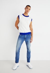 White sleeveless sweater with blue trim, paired with light blue denim jeans and white sneakers. Simple design, casual fit.