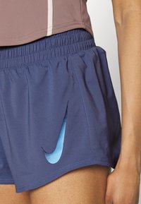 Primer plano de una persona que lleva pantalones cortos deportivos Nike azules con una cinturilla elástica y un logotipo de swoosh de Nike en azul claro en el lado izquierdo.