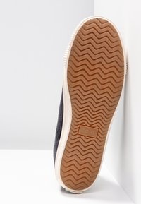 Chaussure grise en toile à enfiler avec une tige texturée, une semelle en caoutchouc présentant un motif en zigzag, et un logo "TOMS" embossé sur le côté.