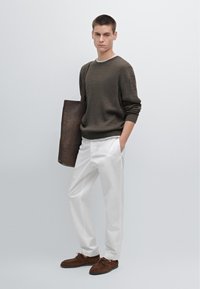 Bruine geribbelde gebreide trui, witte broek, bruine suède schoenen en een donkere leren schoudertas. Model staat tegen een lichte achtergrond.