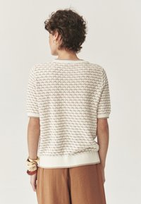 Suéter de punto en blanco roto con un patrón texturizado de diamantes, mangas cortas y dobladillo acanalado. Combinado con pantalones holgados marrones.