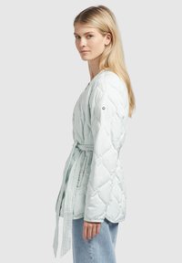 Lichtblauwe gewatteerde jas met een wikkelontwerp, vastgebonden in de taille. Heeft een soepele textuur en schouderknopen. Draagt over lichtblauwe denim.