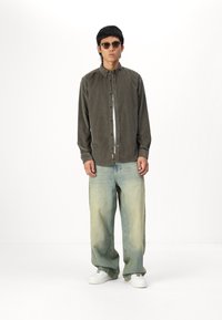 Chemise en velours côtelé vert olive à boutons, jean baggy bleu clair et baskets blanches. Des lunettes de soleil rondes complètent la tenue.