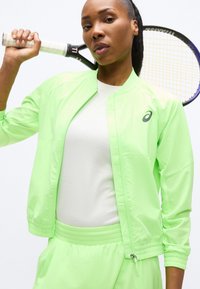 Γυναίκα που κρατά ρακέτα τένις στον ώμο φορώντας αθλητικό μπουφάν και παντελόνι νέον πράσινου χρώματος με λευκό πουκάμισο, μπροστά σε λευκό φόντο.