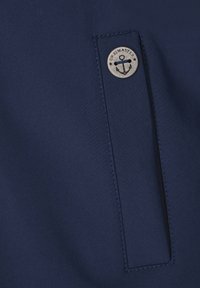 Țesătură de culoare bleumarin, cu o textură netedă, ce prezintă o buzunar și un logo metalic rotund cu un design de ancoră. Detalii cusute vizibile.