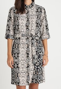 Abito a camicia con stampa a serpente, caratterizzato da un colletto, maniche corte arrotolate e una cintura abbinata. I colori includono beige, nero e bianco.