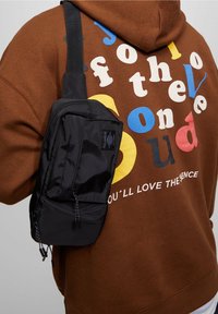 Bolso de lona negro con un compartimento con cremallera, que presenta detalles de cuerda y un parche con el logo. Lleva puesto sobre un sudadera marrón con texto colorido.