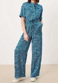 Combinaison bleue à motifs avec manches courtes, taille élastique et jambes larges. Présente un imprimé feuille et une texture de tissu lisse. Portée avec des baskets blanches.