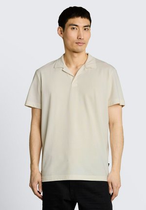 Junger Mann mit kurzen schwarzen Haaren, der ein beiges kurzärmeliges Poloshirt und schwarze Hosen trägt, steht vor einem schlichten hellen Hintergrund.