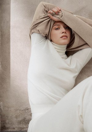 Persoon die een witte coltrui en een beige trui draagt, de trui boven het hoofd tilt, tegen een beige muur met textuur.