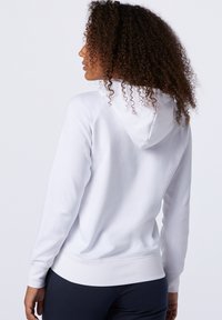 Felpa bianca realizzata in un tessuto morbido, con cappuccio regolabile tramite coulisse, polsini a costine e vita aderente. La vista posteriore mostra un orlo curvato.