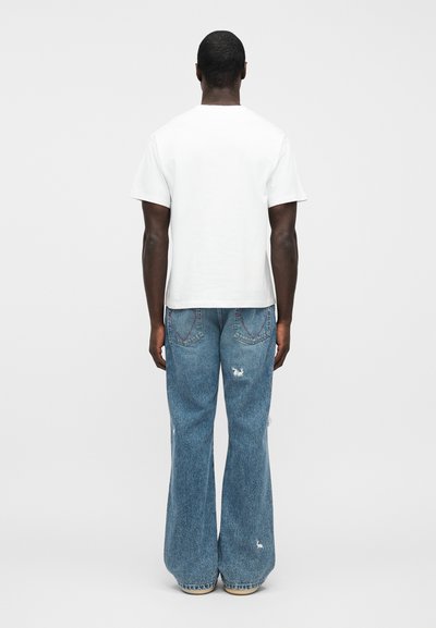 Άνδρας που φοράει λευκό κοντομάνικο μπλουζάκι και φαρδιά μπλε τζιν με μικρές σκισίματα, στέκεται γυρισμένος πλάτη σε απλό φόντο.