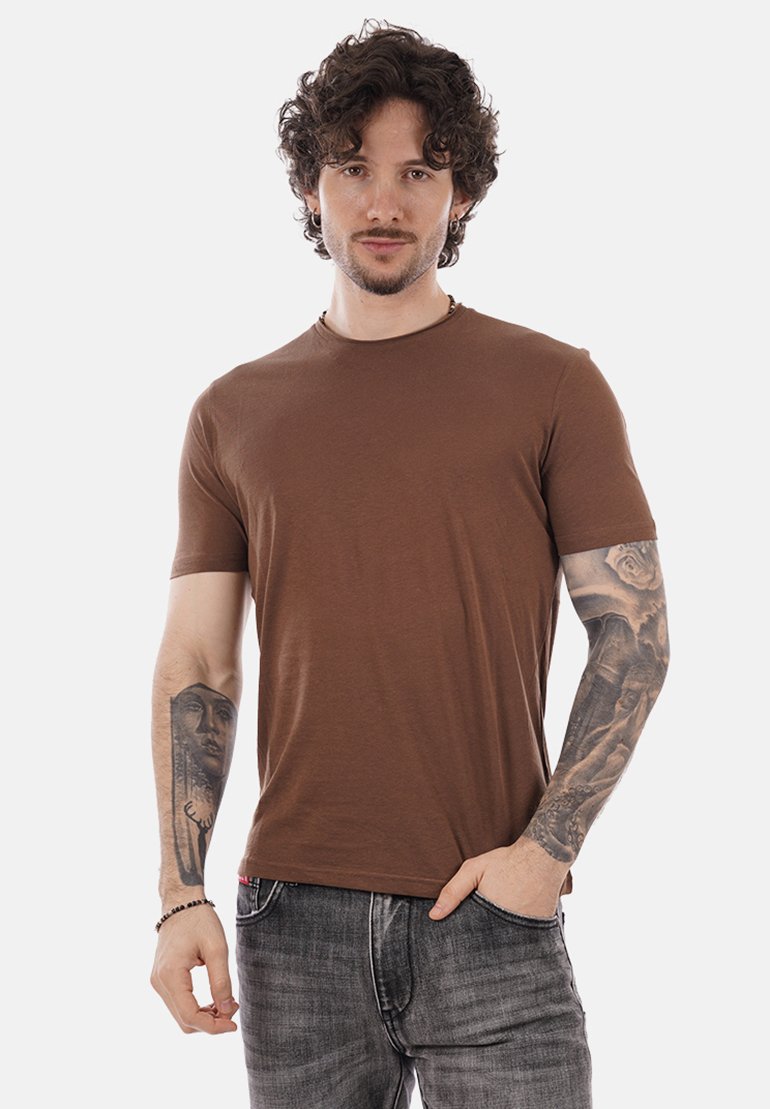 Homme aux cheveux bouclés foncés et barbe, portant un t-shirt marron et un jean gris, montrant des tatouages détaillés sur les deux avant-bras, debout devant un fond blanc.