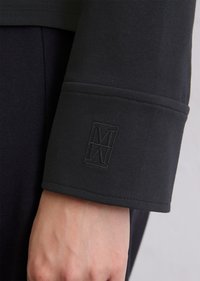 Polsino della camicia nera a maniche lunghe in morbido tessuto con logo rialzato e testurizzato. Il polsino presenta cuciture rinforzate e un taglio dritto.