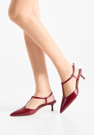 Gambe con décolleté rosse in vernice con cinturino a T, punta appuntita e fibbie piccole, posate su sfondo bianco.