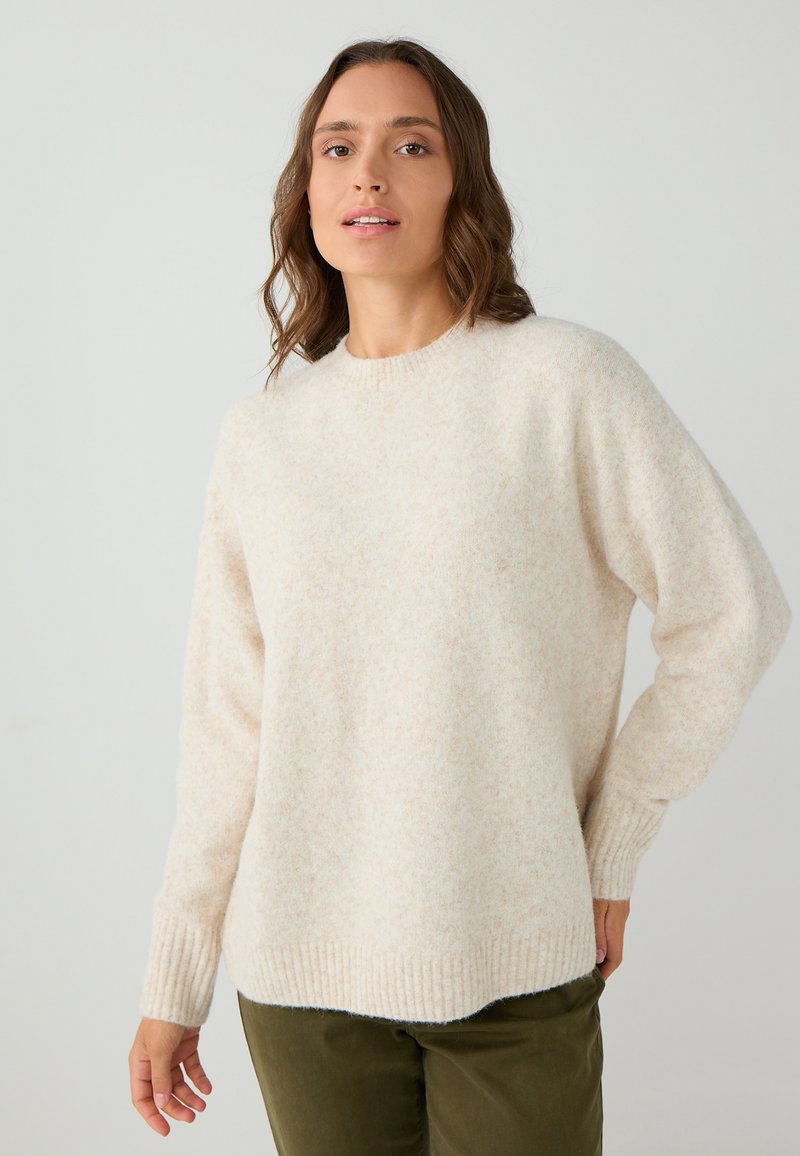 Pull en tricot couleur crème avec une finition texturée, col rond et poignets côtelés. Porté sur un pantalon vert olive.