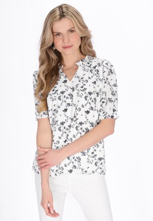 Femeie cu păr blond, ondulat și lung, purtând o bluză albă cu imprimeu floral negru și pantaloni albi, pozând cu brațele încrucișate.