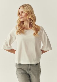 Haut en coton blanc à encolure ronde et manches trois-quarts larges, associé à un jean en denim gris clair. Design minimaliste, texture lisse.