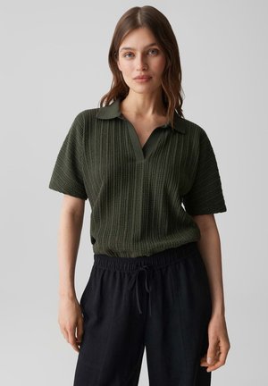 Femme aux cheveux bruns mi-longs portant un haut texturé à manches courtes vert foncé et un pantalon noir à cordon sur un fond uni.