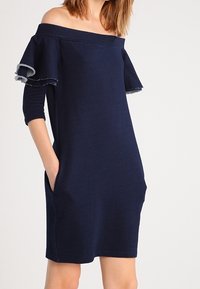 Granatowa sukienka z dekoltem off-the-shoulder, krótkimi rękawami, falbankami i kieszeniami z przodu. Miękki materiał w luźnym kroju.
