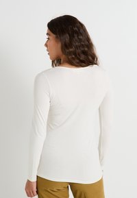 Camiseta de manga larga en color blanco roto, fabricada en una tela suave, con un cuello redondo y un corte ajustado, con una ligera curva en el dobladillo.