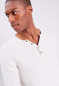 Hellgraues gestricktes Henley-Shirt mit rundem Ausschnitt, drei braunen Knopfakzenten und langen Ärmeln, aus einem weichen, strukturierten Stoff.