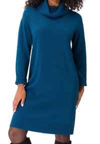 Femme portant une robe en maille bleu sarcelle à manches longues avec des poignets retroussés et un col bénitier, associée à des bottes noires montantes jusqu'aux genoux.