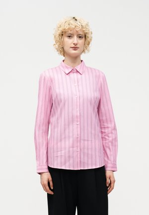 Άτομο με ξανθά, σγουρά μαλλιά που φοράει ροζ πουκάμισο με μακριά μανίκια και λεπτές κάθετες ρίγες, καθώς και μαύρο παντελόνι, στέκεται μπροστά σε λευκό φόντο.