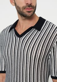 Homme barbu portant un polo en maille rayé noir et blanc à manches courtes avec un col noir et un col en V.