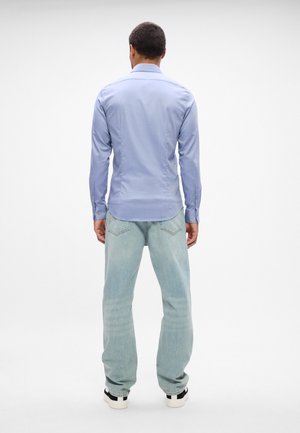 Homme debout, de dos, portant une chemise bleu clair à manches longues, un jean bleu clair et des baskets blanches sur un fond blanc uni.