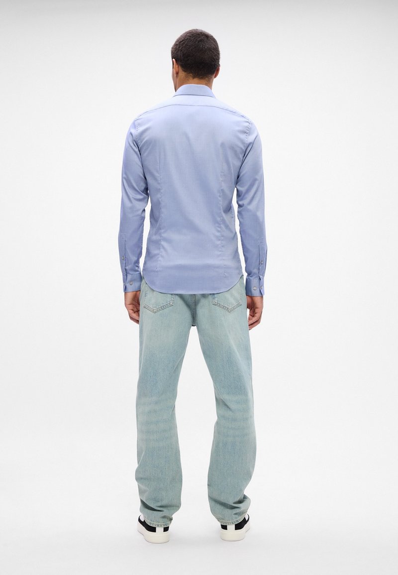 Homme debout, de dos, portant une chemise bleu clair à manches longues, un jean bleu clair et des baskets blanches sur un fond blanc uni.