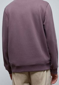 Fioletowy bawełniany sweter z klasycznym okrągłym dekoltem, z ściągaczami przy mankietach i u dołu, prezentowany od tyłu na neutralnym tle.