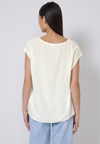Blusa gialla chiaro con maniche corte con dettagli in pizzo, scollatura rotonda e orlo curvo, realizzata in un tessuto morbido e fluido.