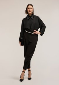 Blusa negra con un cuello tachonado, combinada con pantalones negros de talle alto y tacones negros con tiras. Tela suave con un ajuste relajado.