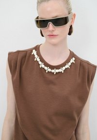 Top sin mangas marrón con tejido texturizado y un cuello decorativo de cuentas blancas y metal. La modelo lleva gafas de sol oscuras y pendientes de aros.