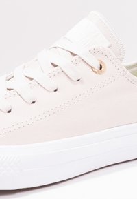 Sneaker en toile beige avec semelle en caoutchouc blanc, dotée d'œillets dorés, de lacets plats et d'une étiquette sur la languette texturée.