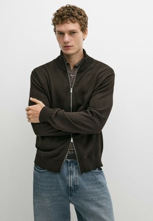 Junger Mann mit lockigem Haar, der einen schwarzen Zip-Up-Pullover über einem gestreiften Hemd und blauen Jeans trägt, mit verschränkten Armen stehend.