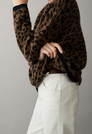 Osoba w brązowo-czarnym swetrze w panterkę z czarnymi mankietami i białych spodniach, trzymająca brzeg swetra jedną ręką.