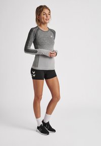 Kvinna stående klädd i en grå långärmad träningsöverdel, svarta shorts, vita strumpor och svarta löparskor mot en enkel ljus bakgrund.