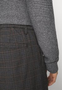 Šedý svetr s texturou a žebrovanými rukávy, doplněný tmavými károvanými kalhotami s modrými a hnědými linkami a detailem zadní kapsy.