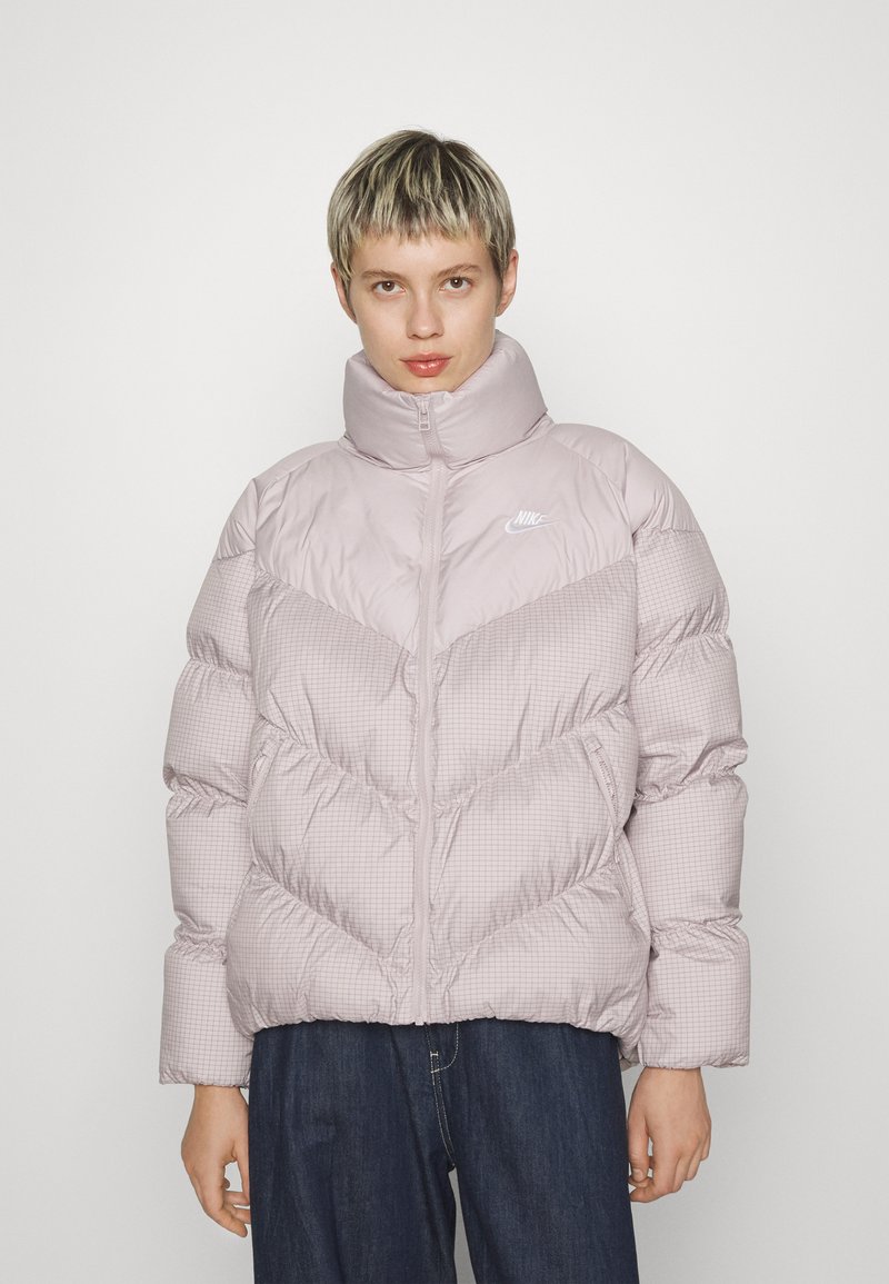 Veste en duvet rose clair avec un col haut, un design en chevron et une texture côtelée. Elle possède une fermeture éclair et des détails de logo sur la poitrine.