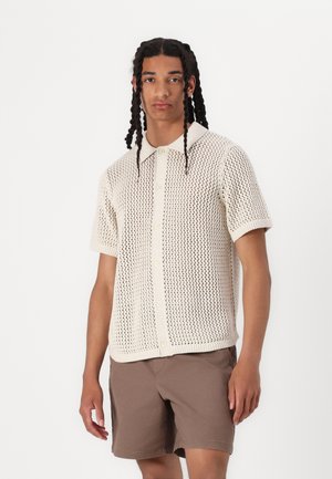 Maglietta lavorata a maglia color crema con chiusura a bottoni sul davanti, maniche corte e un design a trama aperta e testurizzata. Abbinata a pantaloni corti marroni.