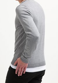 Grå stickad tröja med långa ärmar och räfflade manchetter, med en enkel design och strukturerad tyg, lagd över en vit skjorta.