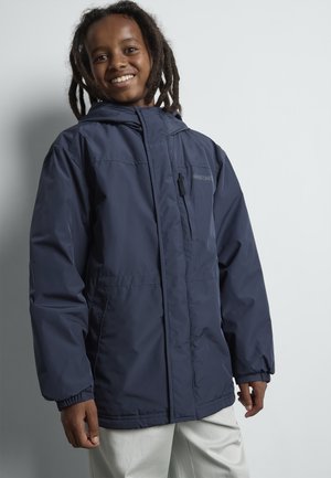 Smiling child with dreadlocks wearing a navy hooded jacket and light-colored pants, standing against a plain light background.